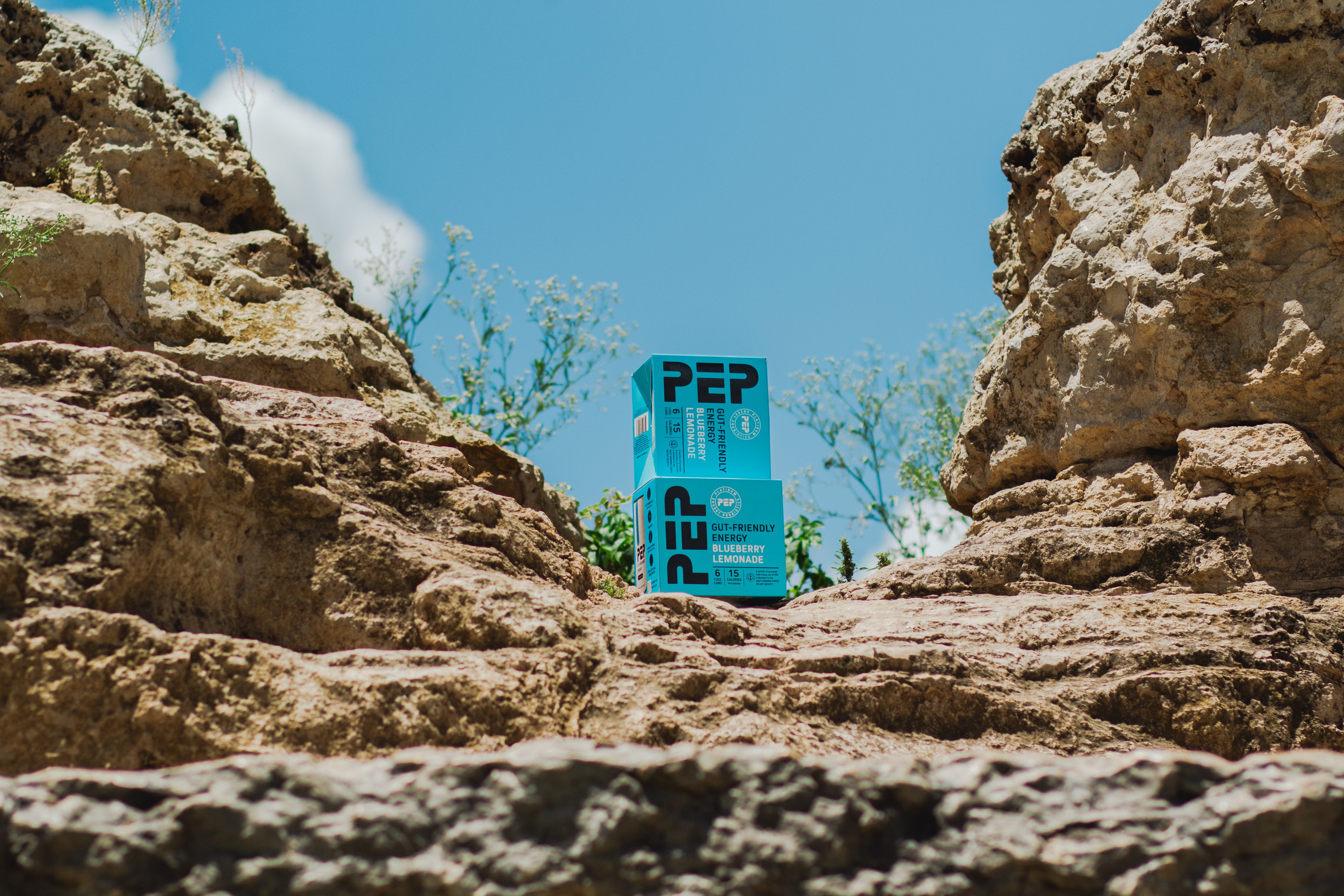 Two PEP Blueberry Lemonade 6-pack boxes on a rocky terrain with a clear blue sky.